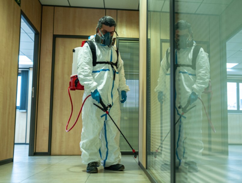 man in a plastic suit with a gas mask sprays pesticide along the base of a wall in an office hallway