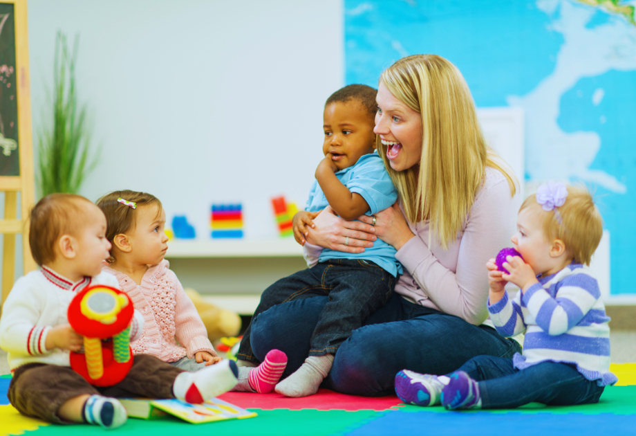 a woman smiles and plays with toddlers in a play area